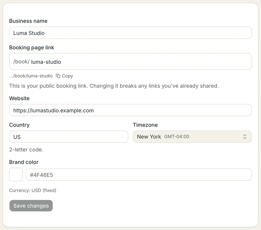 The Business details form: the salon's name, its public booking-page link shown as /book/ luma-studio with a preview and a Copy control beneath it, a website address, a two-letter country code, a timezone picker reading New York with its GMT offset, an unset brand-colour field showing an example hex code beside a blank swatch, and a line stating the currency is USD and fixed.